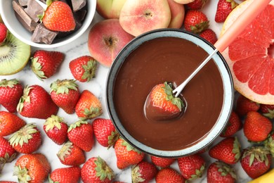 Photo of Fondue fork with strawberry in bowl of melted chocolate surrounded by other fruits on white marble table, flat lay