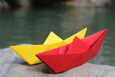Photo of Beautiful yellow and red paper boats on stone near water outdoors, closeup