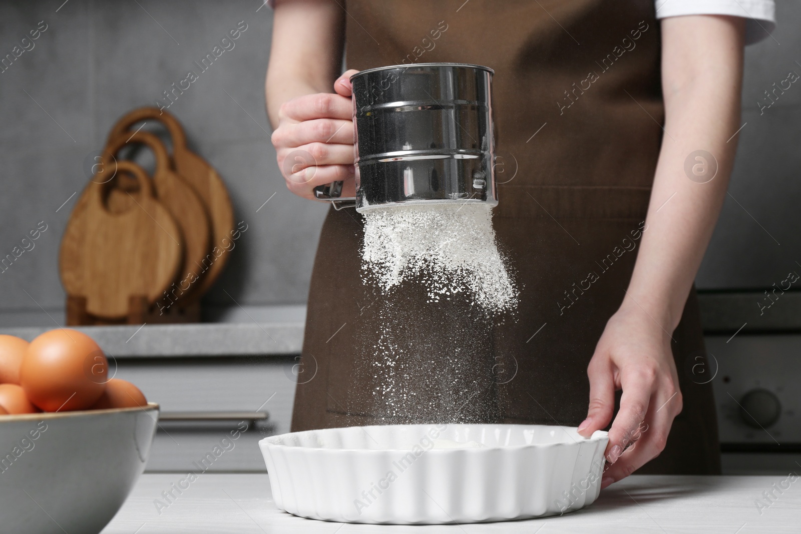 Photo of Woman sieving flour into baking dish at table in kitchen, closeup