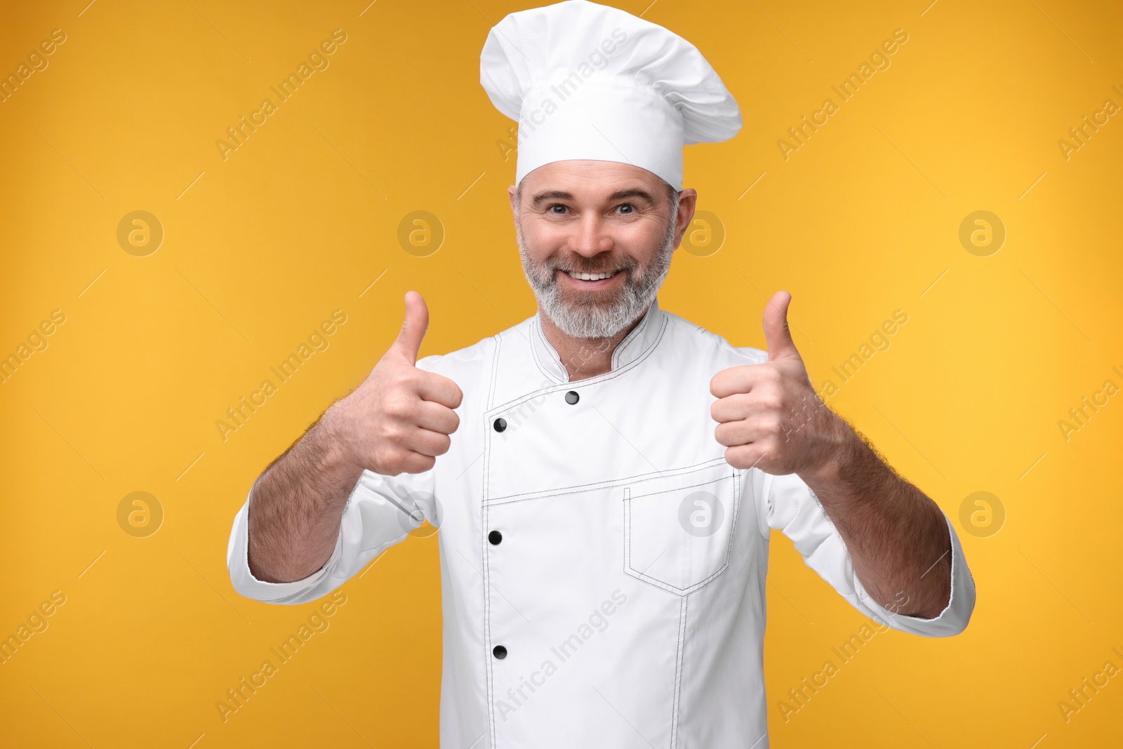 Photo of Happy chef in uniform showing thumbs up on orange background