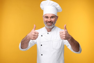 Happy chef in uniform showing thumbs up on orange background