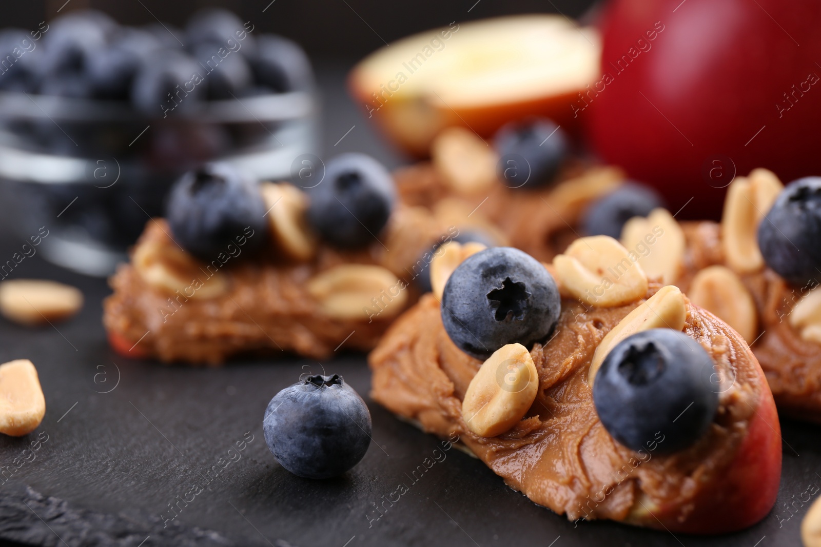 Photo of Pieces of fresh apple with peanut butter and blueberries on dark board, closeup