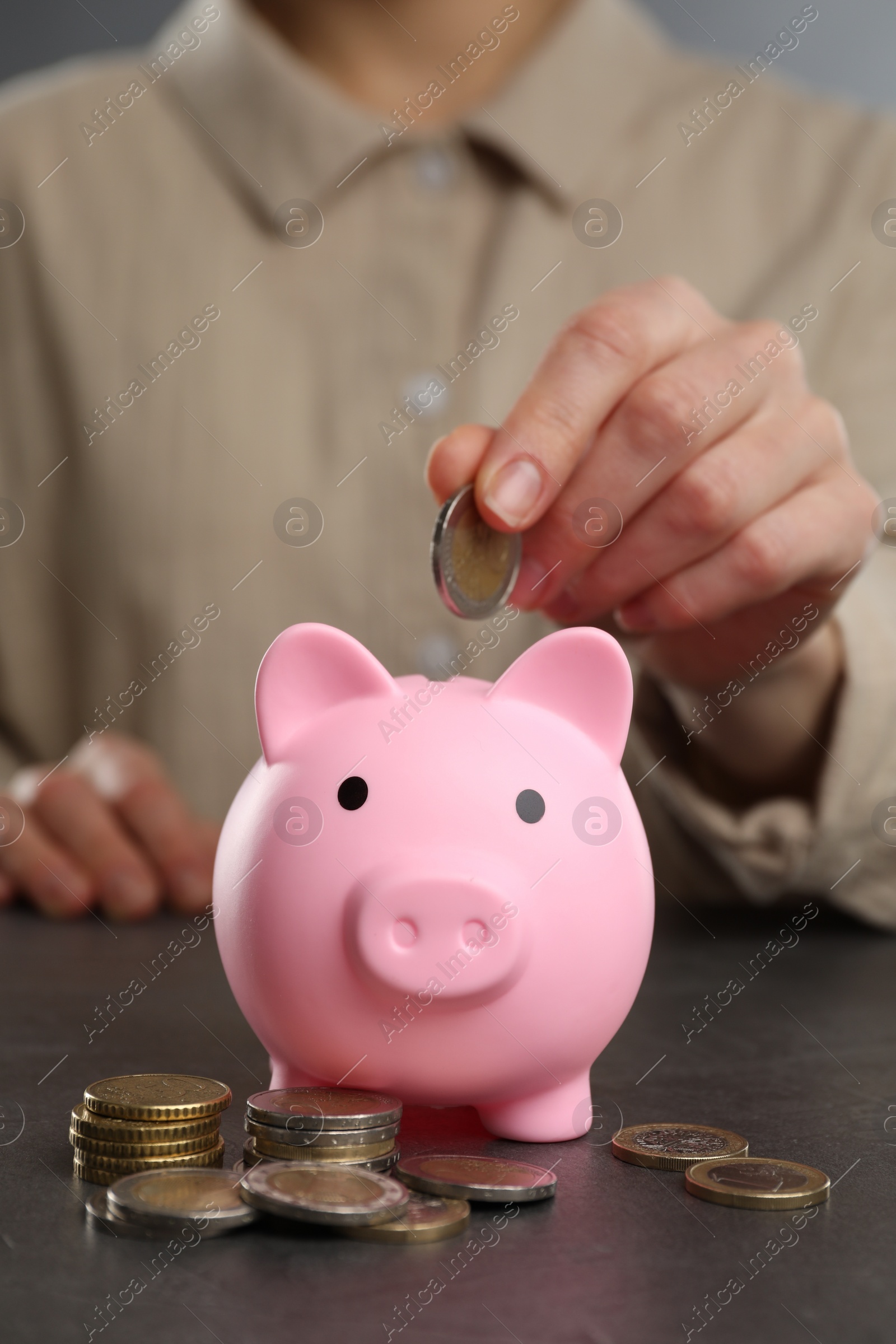 Photo of Woman putting coin into pink piggy bank at black table, closeup