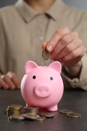 Photo of Woman putting coin into pink piggy bank at black table, closeup