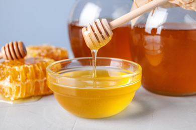 Photo of Dripping tasty honey from dipper into bowl on light grey table, closeup