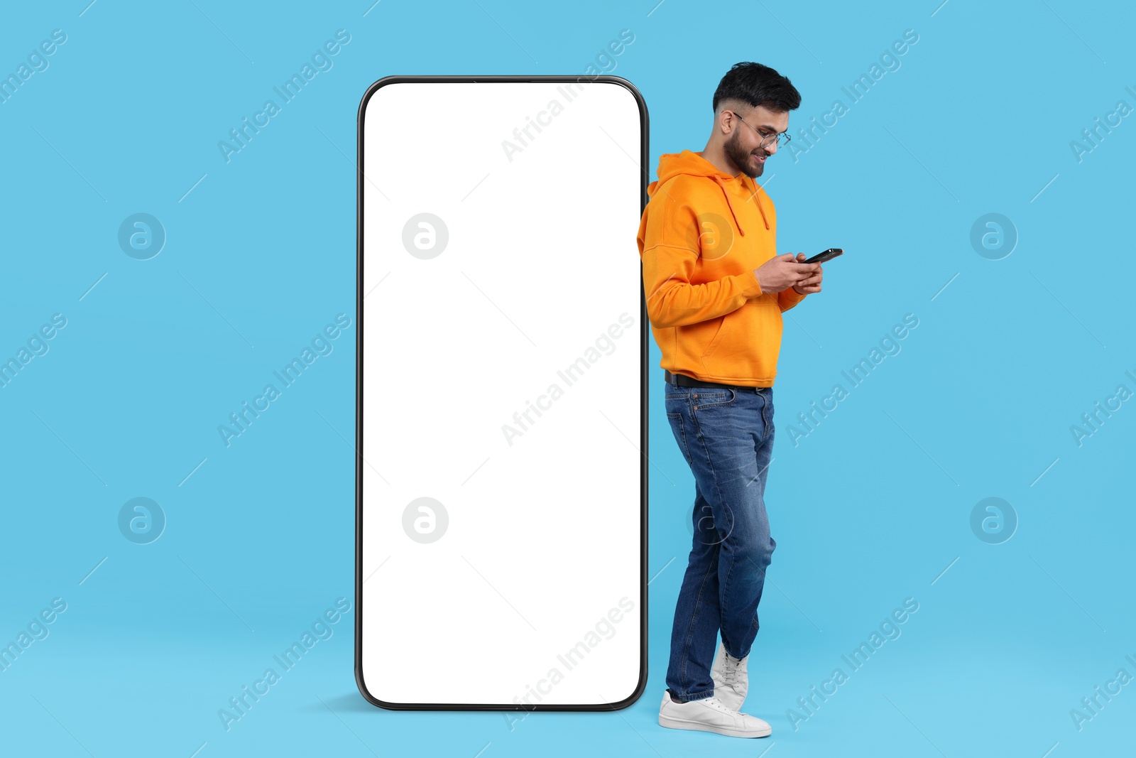 Image of Man with mobile phone standing near huge device with empty screen on light blue background. Mockup for design