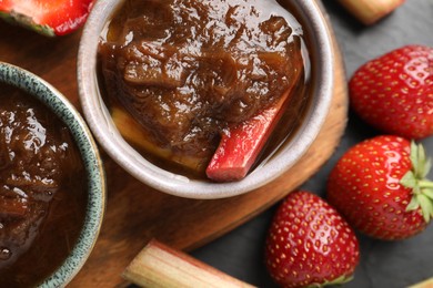 Tasty rhubarb jam in bowls, stems and strawberries on dark table, flat lay