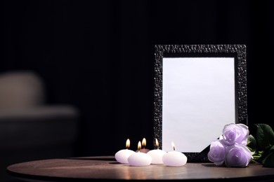 Funeral photo frame with black ribbon, violet roses and burning candles on wooden table indoors. Space for text