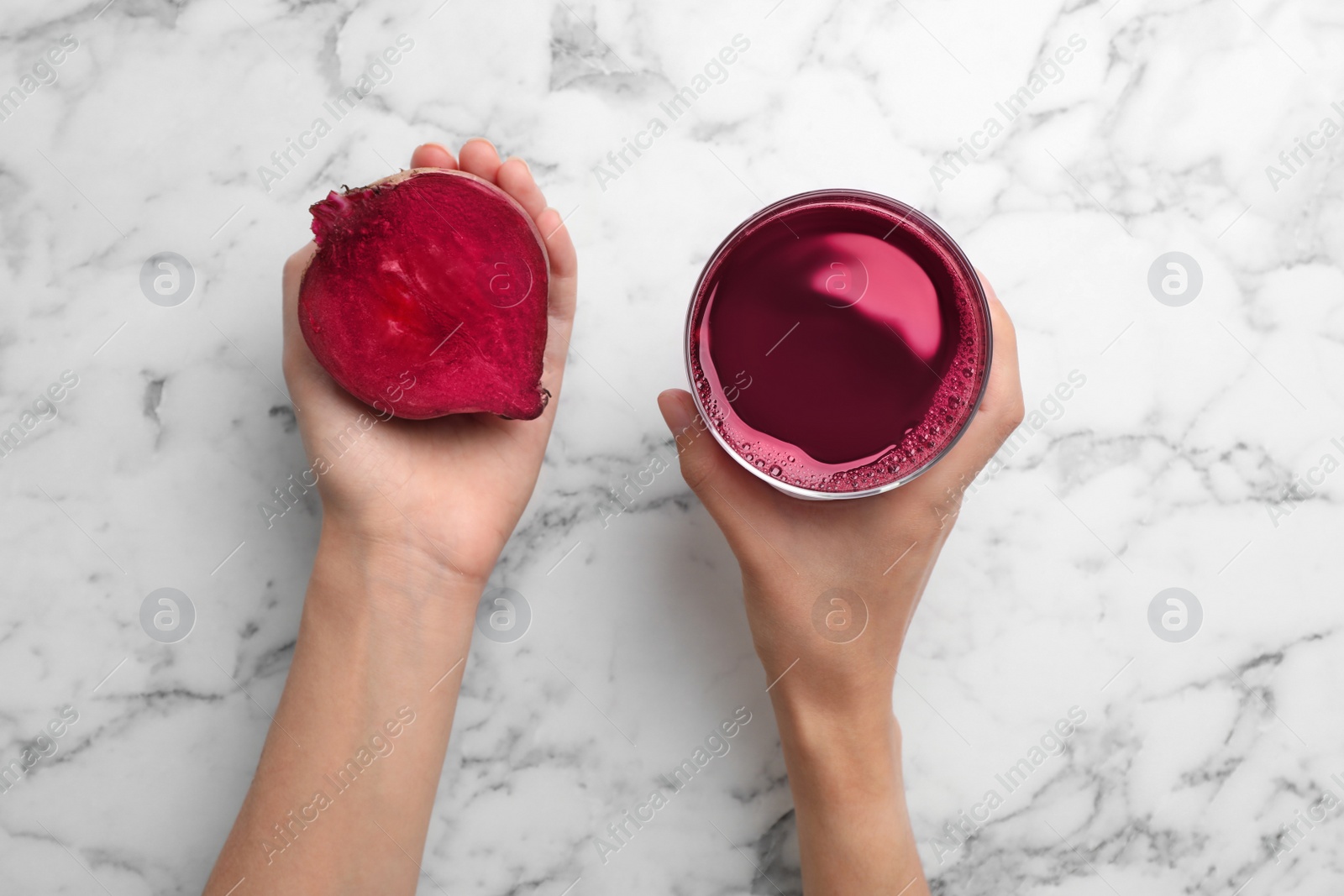 Photo of Woman holding glass with fresh beet juice and half of vegetable on marble table, top view