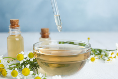 Photo of Chamomile essential oil dripping from pipette into bowl on white wooden table, closeup