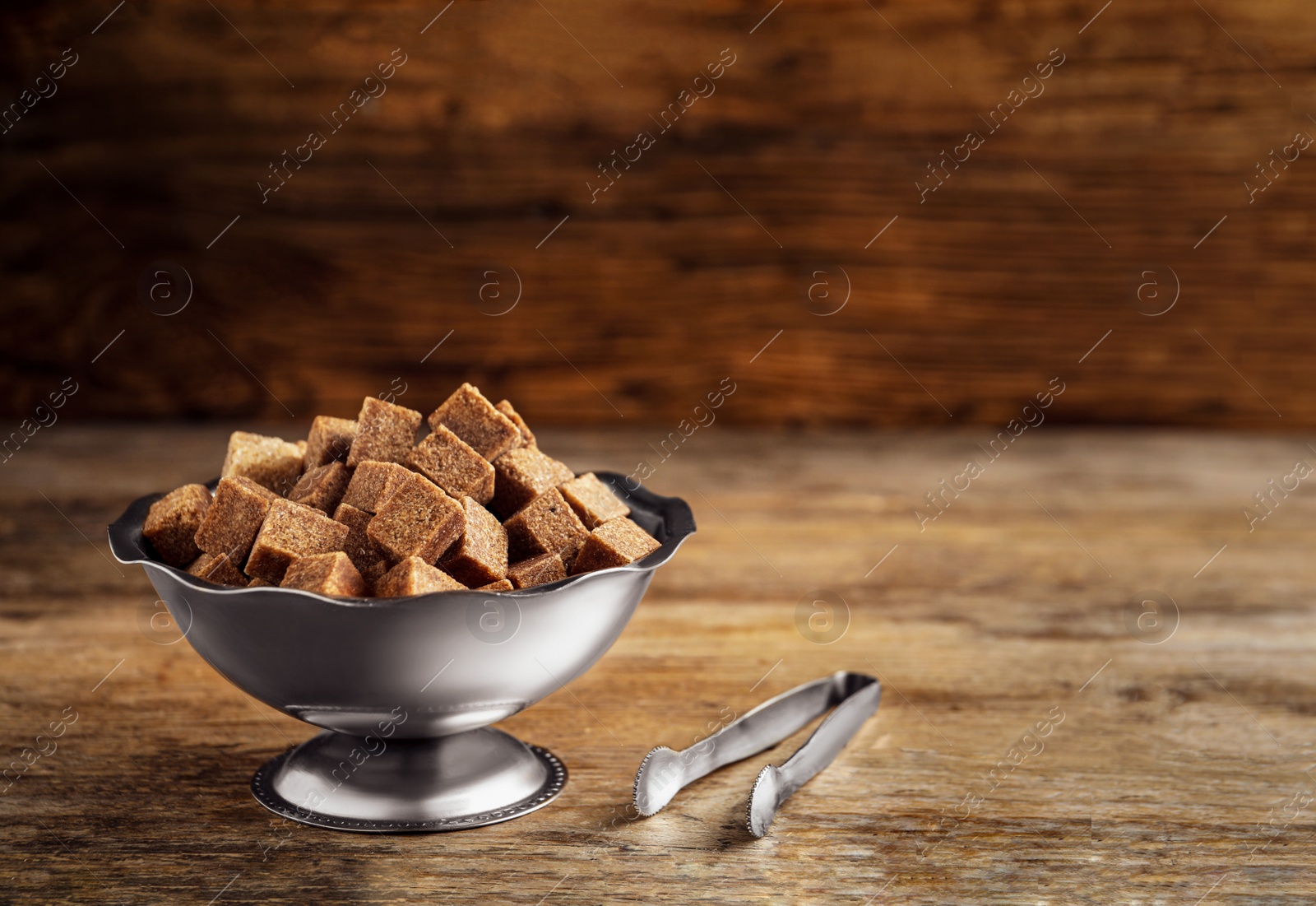 Photo of Brown sugar cubes in metal bowl and tongs on wooden table. Space for text