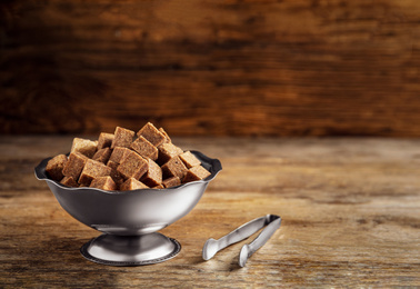Photo of Brown sugar cubes in metal bowl and tongs on wooden table. Space for text
