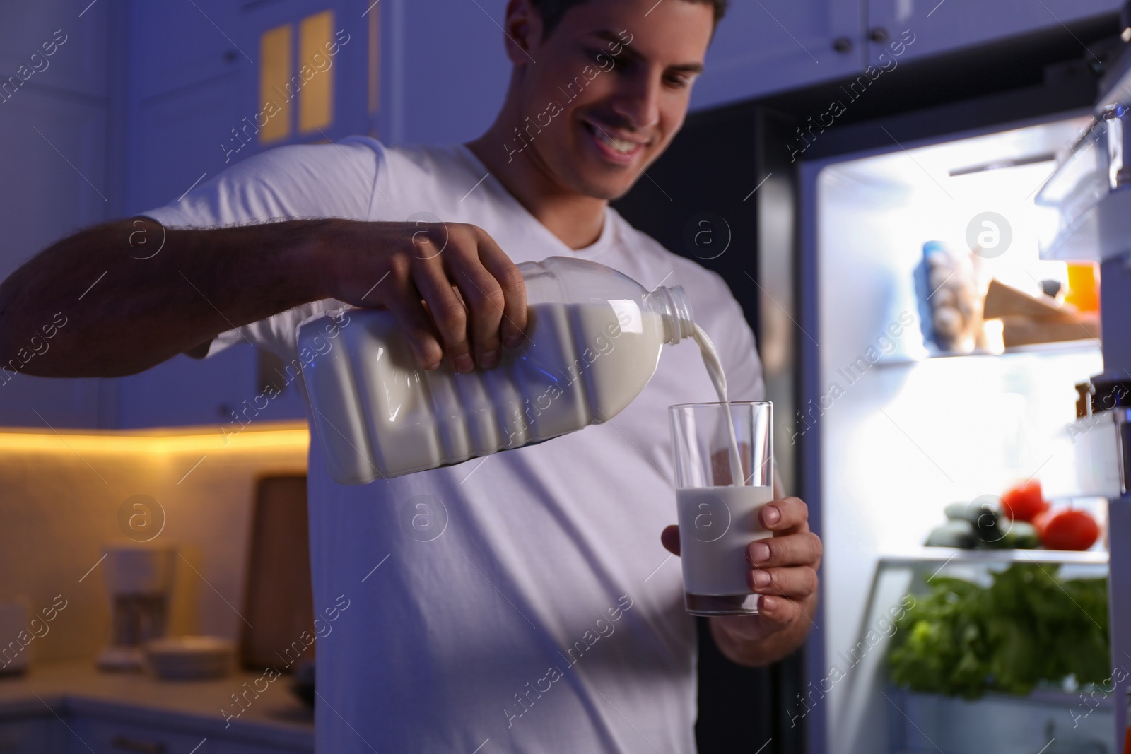 Photo of Man pouring milk from gallon bottle into glass near refrigerator in kitchen at night