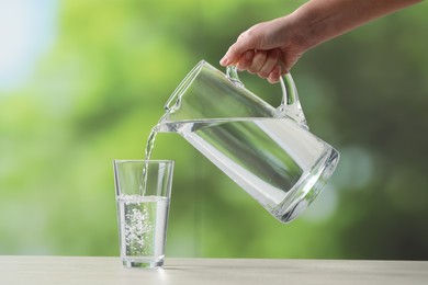 Woman pouring fresh water from jug into glass at white table against blurred green background, closeup