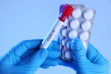 Scientist holding test tube with blood sample and pills on blue background, closeup