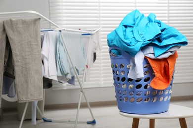 Photo of Plastic laundry basket overfilled with clothes near drying rack on white stool indoors. Space for text