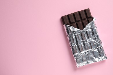 Photo of Delicious dark chocolate bar wrapped in foil on pink background, top view. Space for text