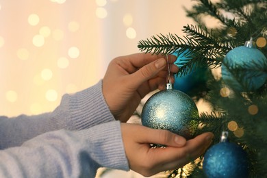 Photo of Woman decorating Christmas tree with light blue festive ball on blurred background, closeup