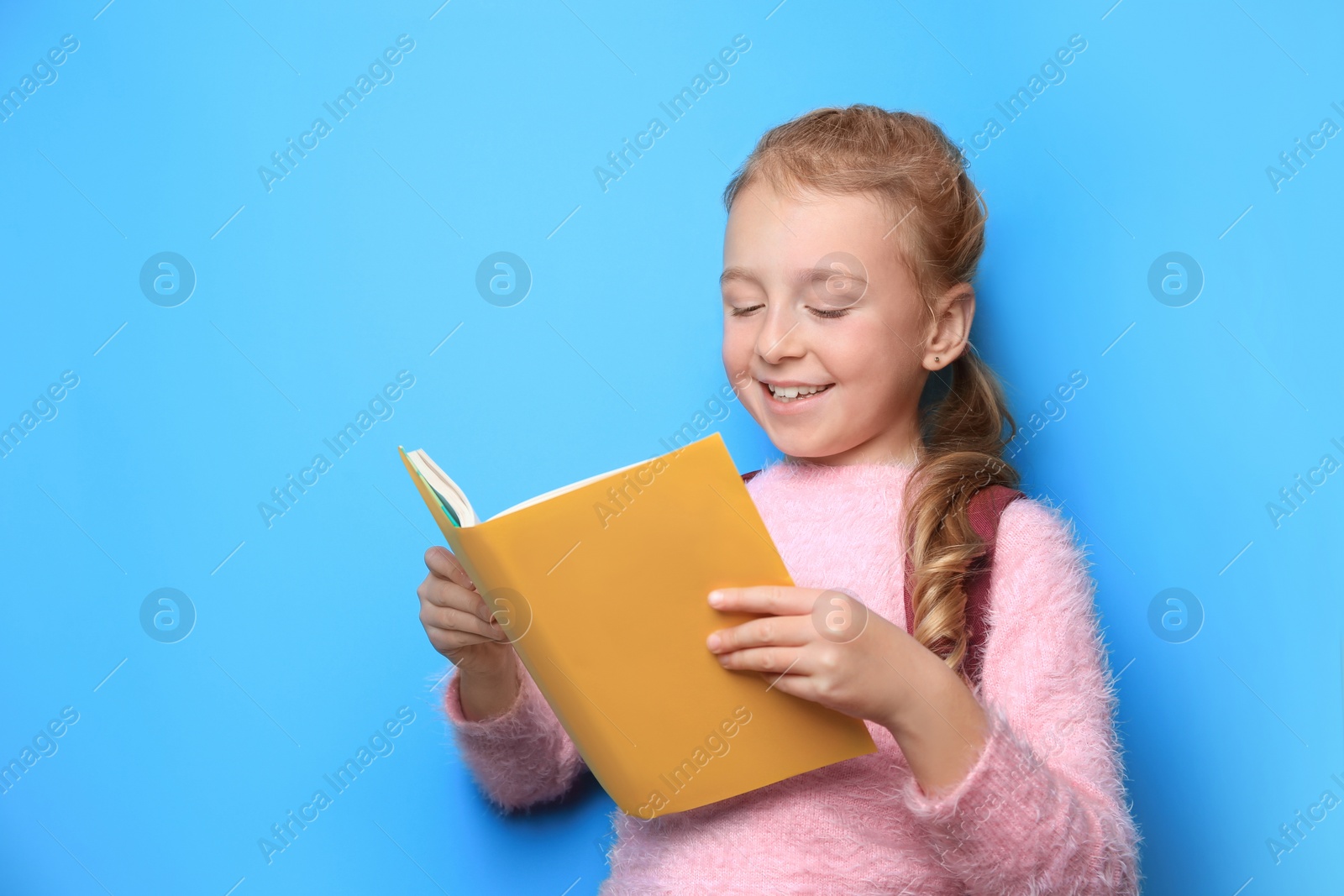 Photo of Happy little girl with backpack reading book on light blue background