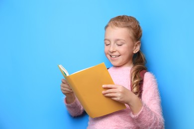 Photo of Happy little girl with backpack reading book on light blue background