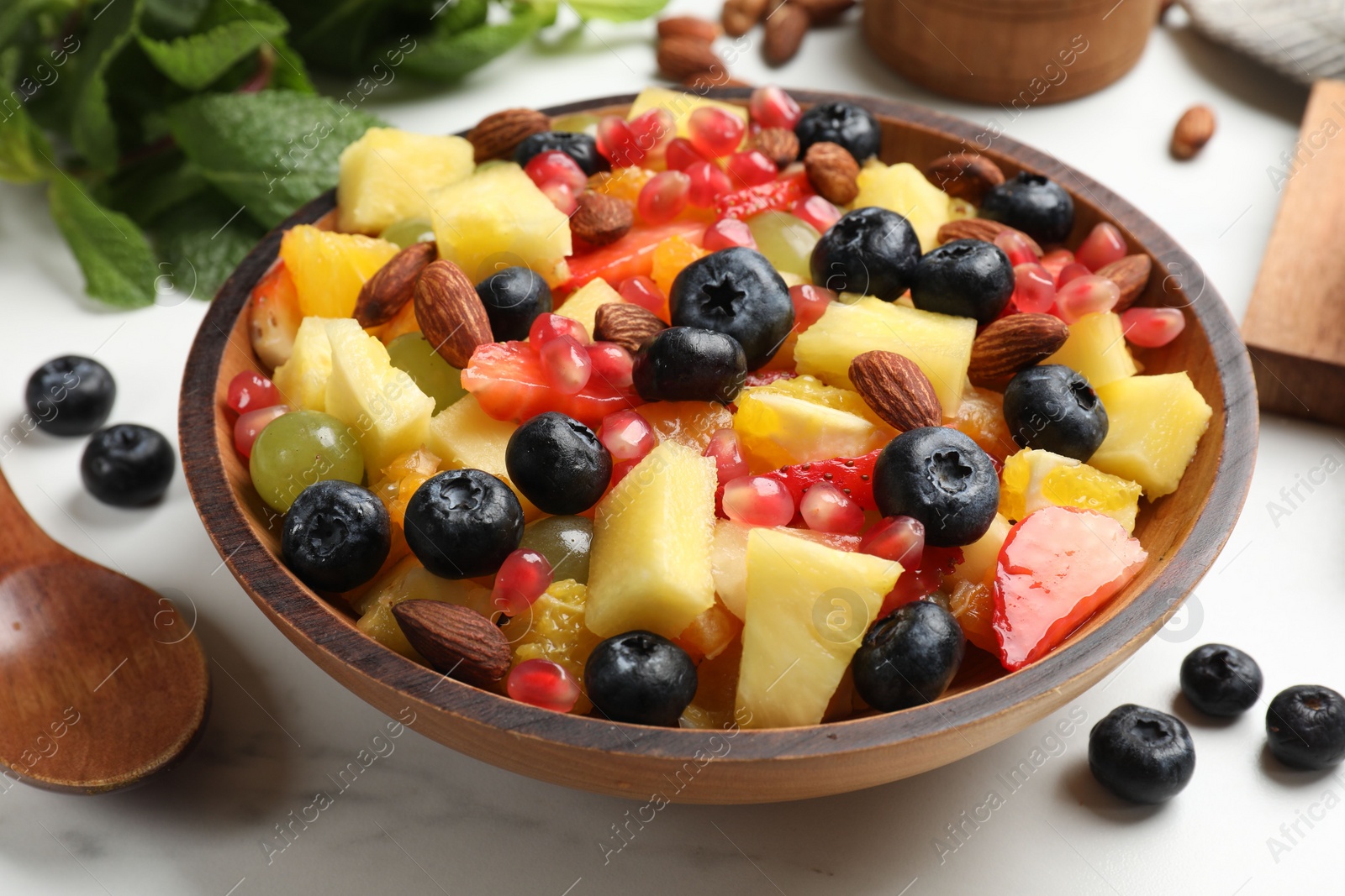 Photo of Delicious fruit salad in bowl, berries, fresh mint and nuts on white marble table, closeup