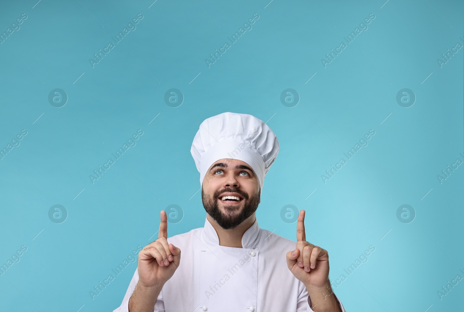 Photo of Happy young chef in uniform pointing at something on light blue background. Space for text