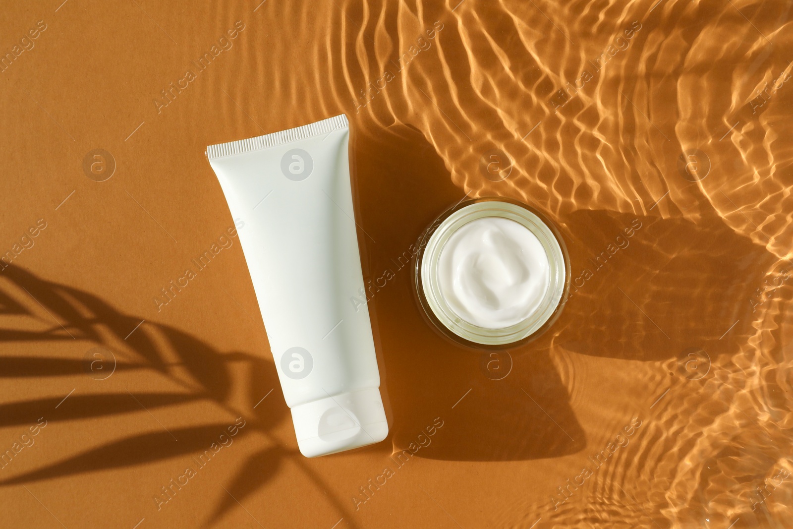 Photo of Tube and jar with moisturizing cream in water on orange background, top view