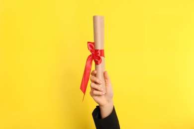 Photo of Student holding rolled diploma with red ribbon on yellow background, closeup