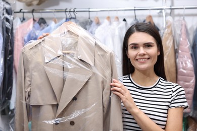 Photo of Dry-cleaning service. Happy woman holding hanger with coat in plastic bag indoors