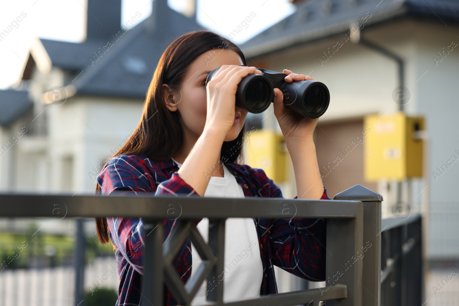 Photo of Concept of private life. Curious young woman with binoculars spying on neighbours over fence outdoors