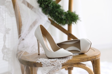 Photo of Pair of white high heel shoes, veil and wreath on wooden chair indoors. Dressing for wedding