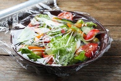 Photo of Bowl of fresh salad with plastic food wrap on wooden table, closeup