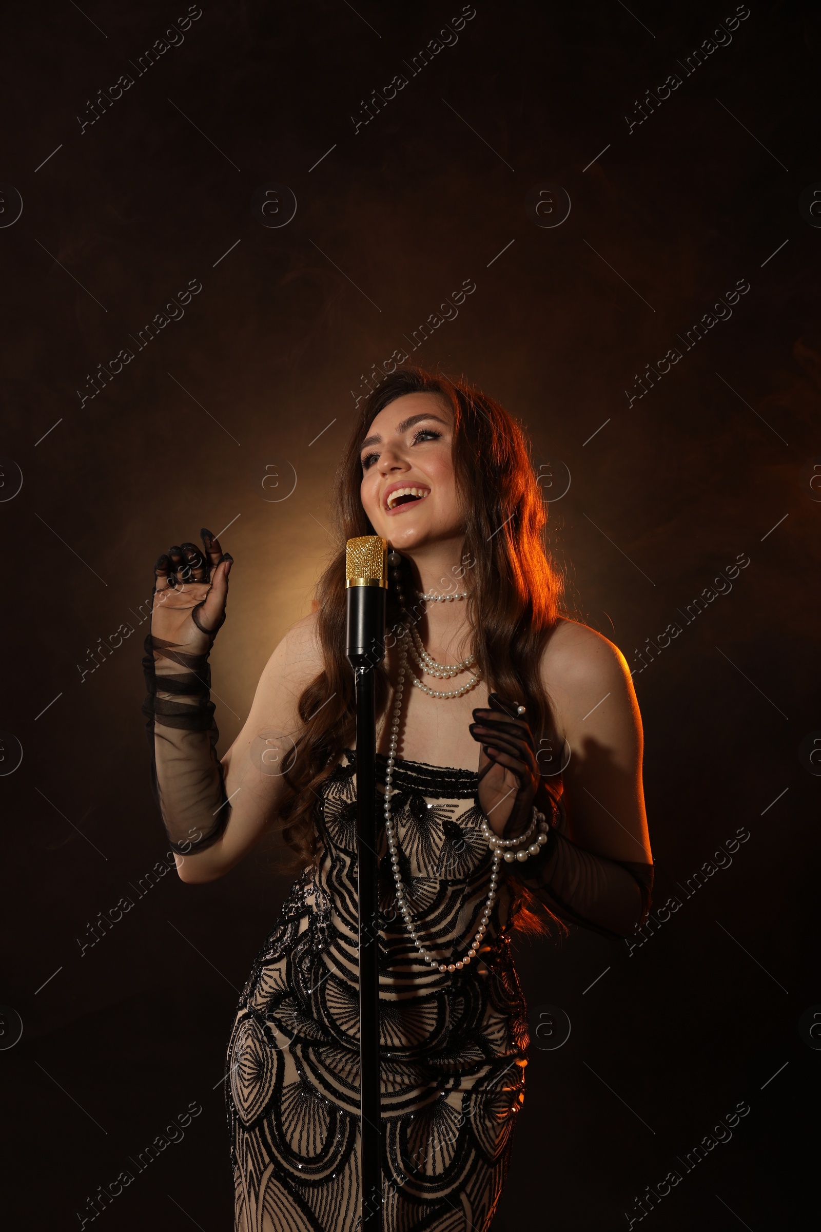 Photo of Beautiful young woman in stylish dress with microphone singing on dark background with smoke