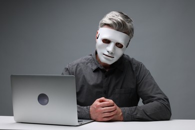 Man in mask sitting at white table with laptop against grey background