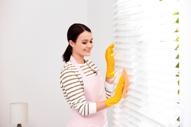 Photo of Young chambermaid wiping dust from blinds in room