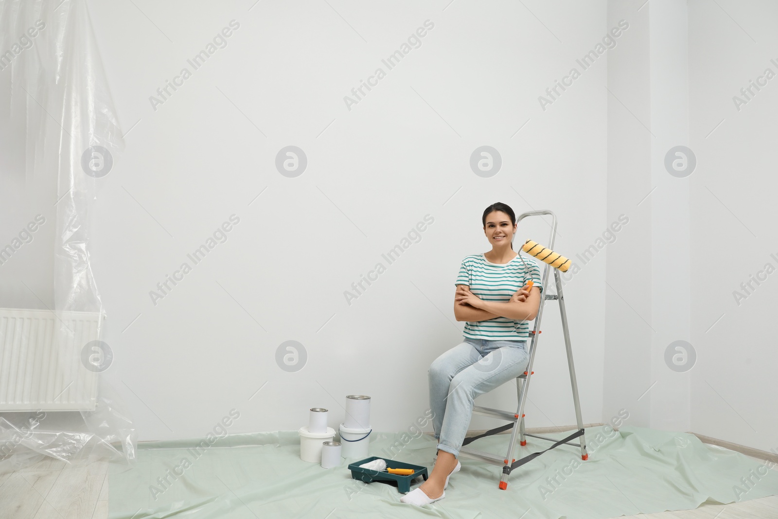 Photo of Woman with paint roller brush sitting on metallic folding ladder indoors, space for text