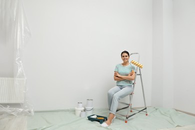 Photo of Woman with paint roller brush sitting on metallic folding ladder indoors, space for text