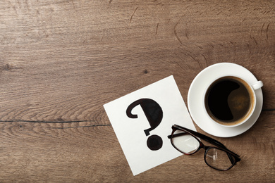 Photo of Note with question mark, eyeglasses and cup of coffee on wooden table, flat lay. Space for text