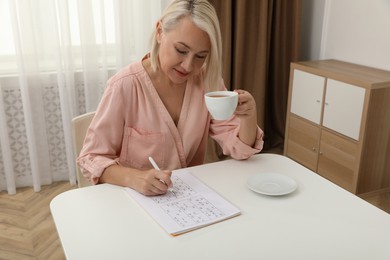 Photo of Middle aged woman with cup of drink solving sudoku puzzle at table indoors