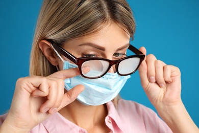 Photo of Woman wiping foggy glasses caused by wearing disposable mask on blue background, closeup. Protective measure during coronavirus pandemic