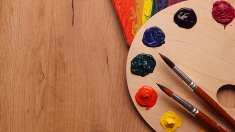 Photo of Artist's palette with samples of colorful paints and brushes on wooden table, flat lay. Space for text