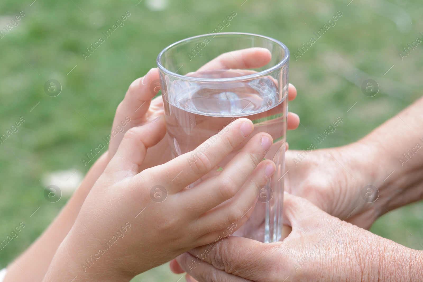 Photo of Child giving glass of water to elderly woman outdoors, closeup