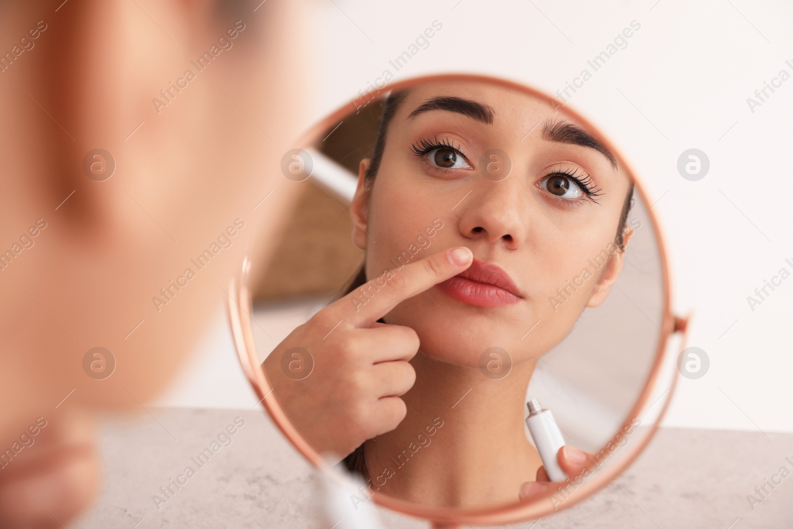 Photo of Woman with herpes touching lips in front of mirror against light background