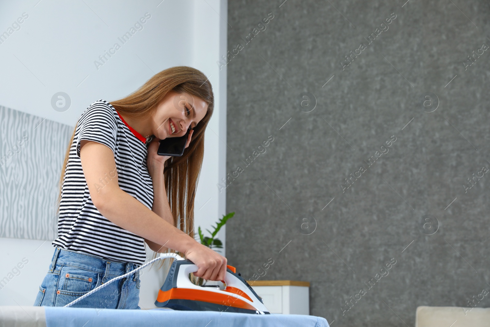 Photo of Happy woman talking on phone while ironing clothes at home. Space for text