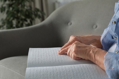 Photo of Blind senior person reading book written in Braille on sofa indoors, closeup. Space for text