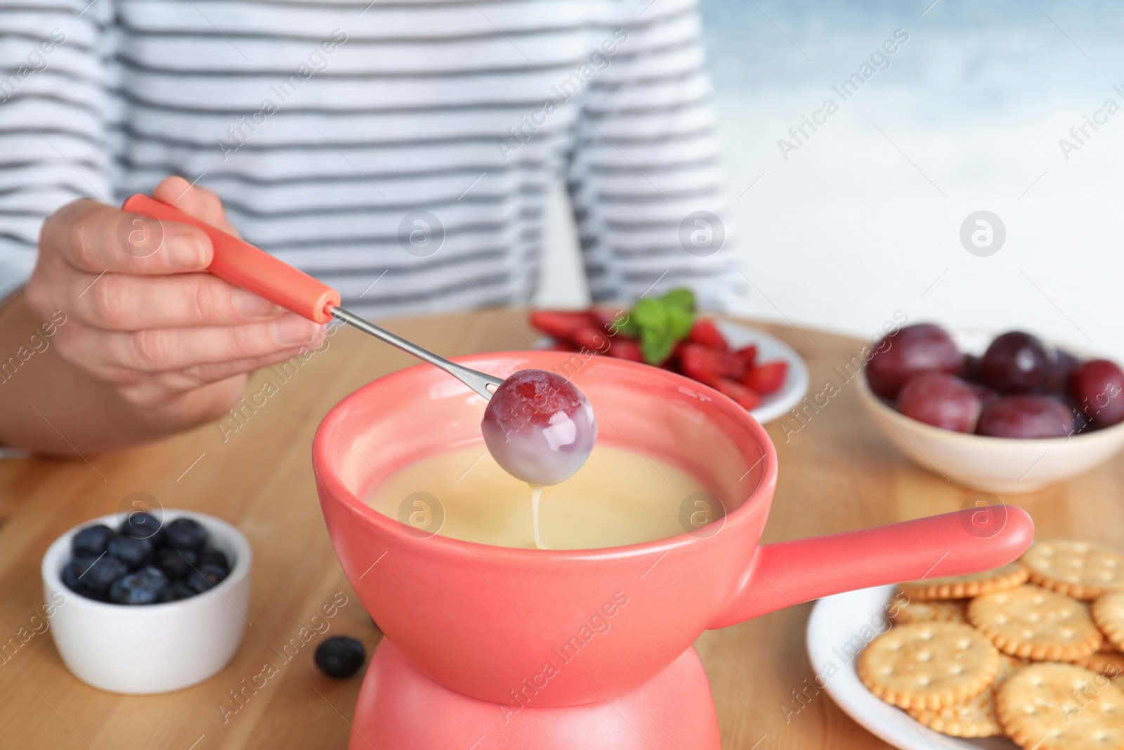 Photo of Woman dipping grape into pot with white chocolate fondue at table, closeup