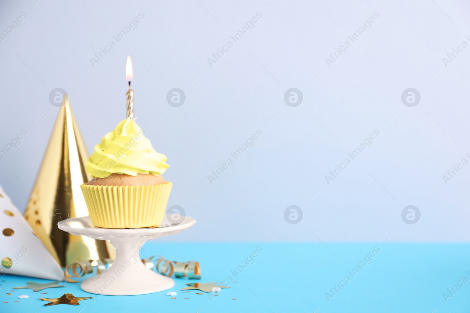 Photo of Delicious birthday cupcake with burning candle and party decor on light blue table against grey background, space for text