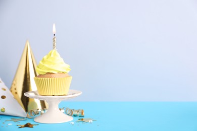 Delicious birthday cupcake with burning candle and party decor on light blue table against grey background, space for text