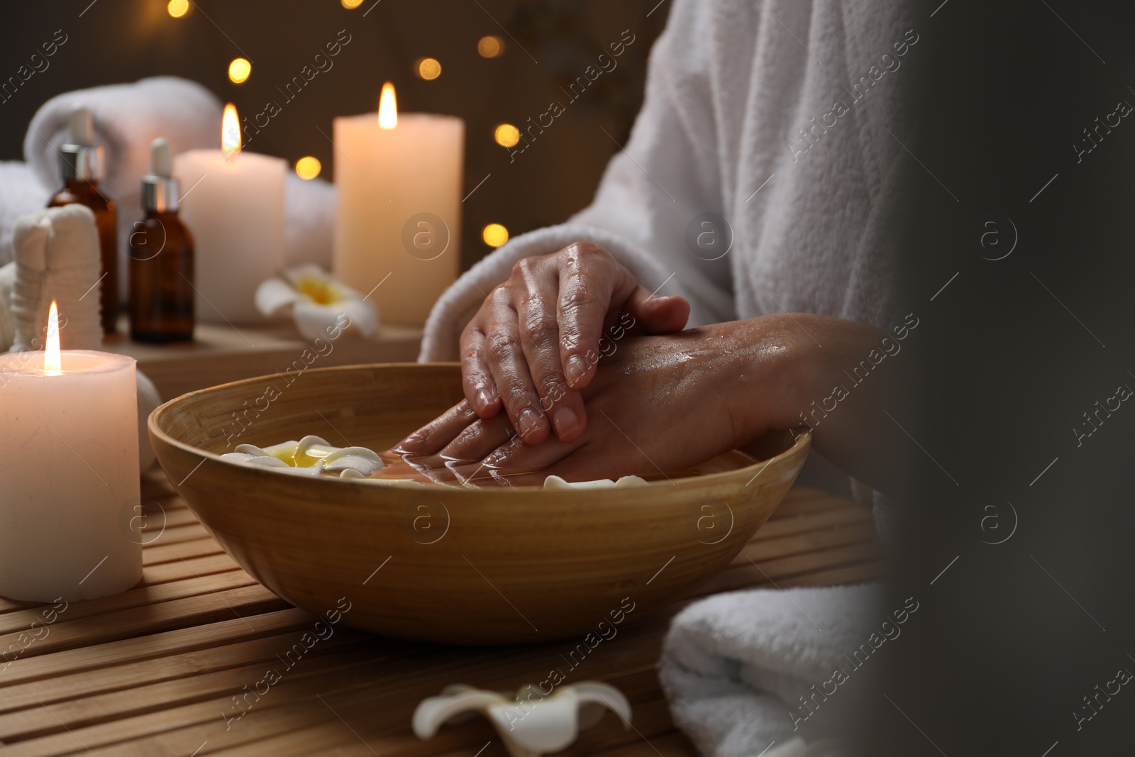 Photo of Woman soaking her hands in bowl of water and flowers at wooden table, closeup. Spa treatment