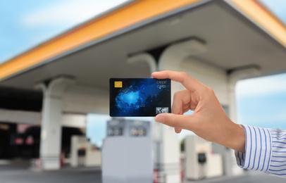 Image of Woman holding credit card against blurred gas station, closeup. Cashless payment